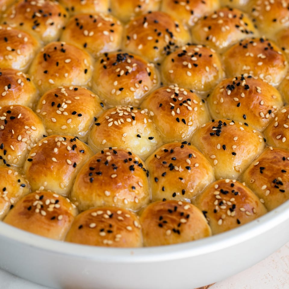 Close-up of golden-brown baked goods, possibly pastries or bread rolls, topped with sesame and black seeds.