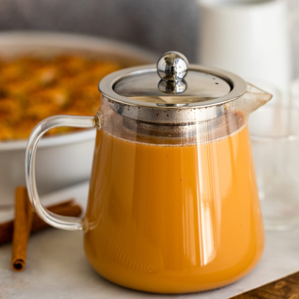 Glass teapot filled with orange chai tea, with cinnamon sticks and a pastry in the background.