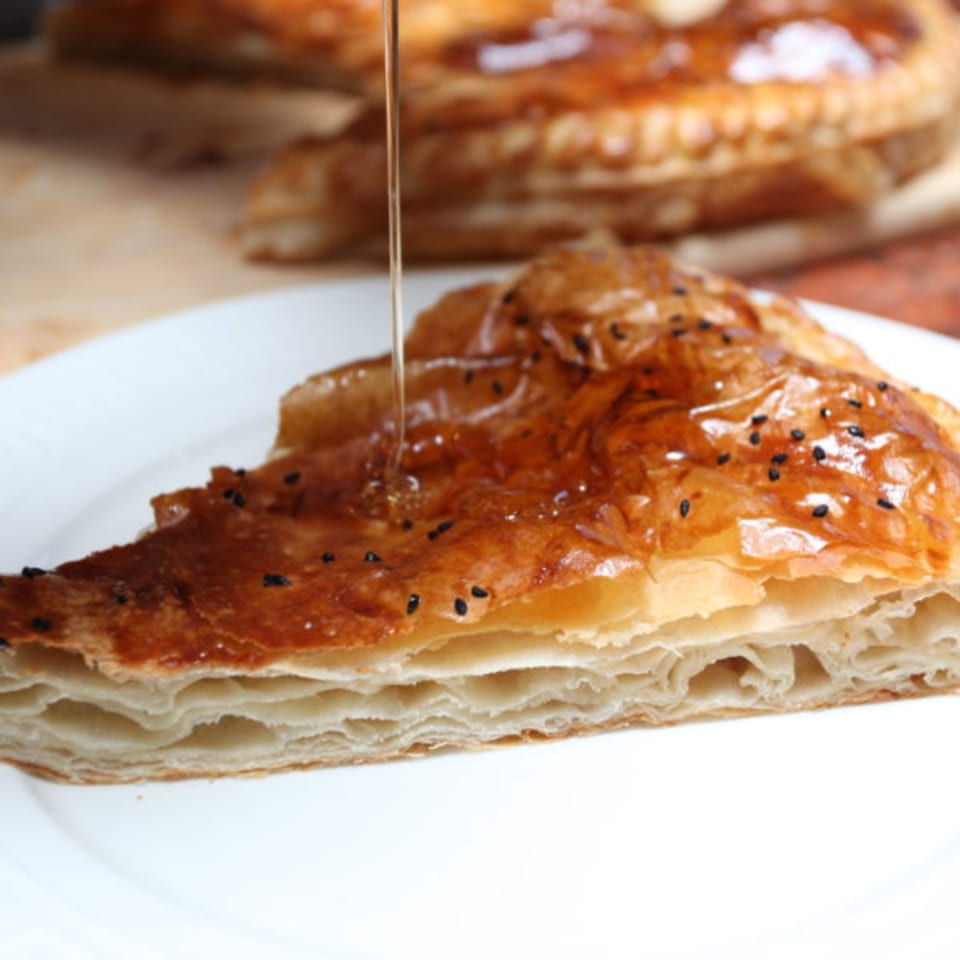 A slice of layered pastry with black sesame seeds, drizzled with honey on a white plate.